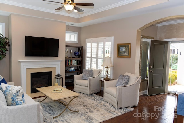 living area featuring arched walkways, crown molding, a glass covered fireplace, ceiling fan, and wood finished floors