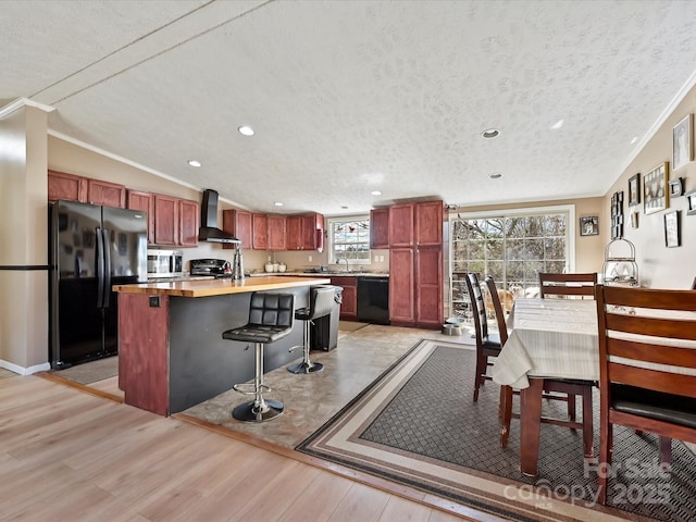 kitchen featuring vaulted ceiling, wall chimney range hood, light wood-type flooring, reddish brown cabinets, and black appliances