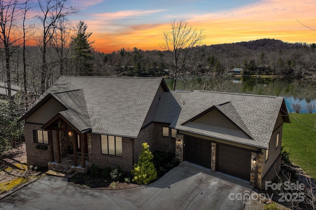 view of front of home with a water view, driveway, a shingled roof, a garage, and brick siding