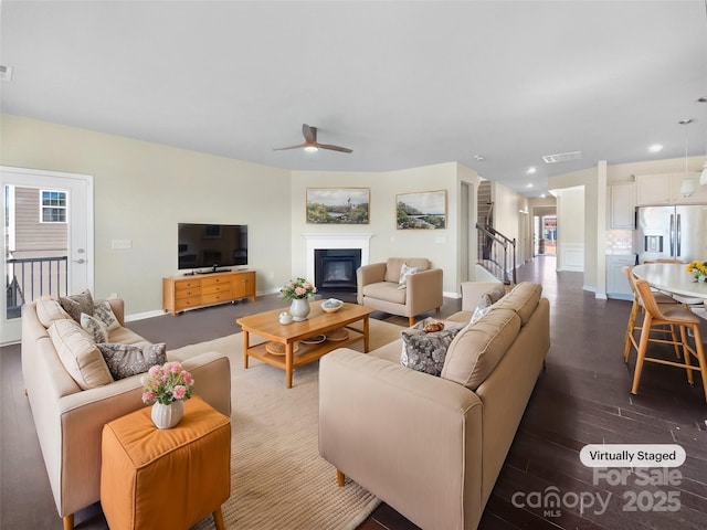 living area featuring dark wood-style floors, baseboards, visible vents, a ceiling fan, and a glass covered fireplace