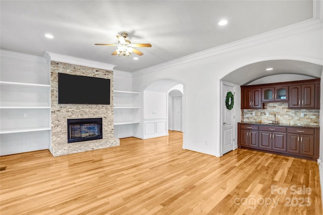 unfurnished living room featuring built in features, ceiling fan, light wood-style floors, crown molding, and a large fireplace