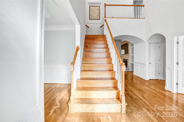 staircase featuring wood finished floors, a wainscoted wall, a stone fireplace, crown molding, and a decorative wall