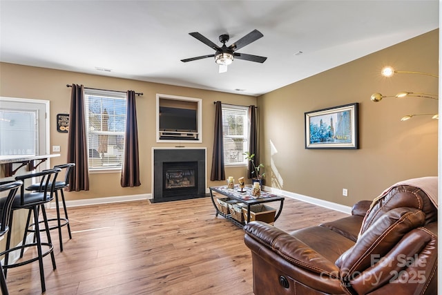 living area with light wood-style flooring, a fireplace with flush hearth, a ceiling fan, and baseboards