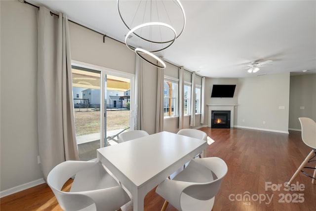 dining space featuring a glass covered fireplace, ceiling fan with notable chandelier, wood finished floors, and baseboards