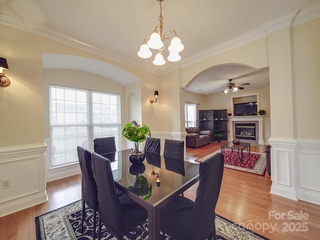 dining room featuring ceiling fan with notable chandelier, wood finished floors, wainscoting, a tiled fireplace, and crown molding