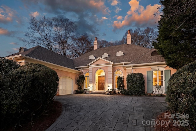 view of front facade with stucco siding, an attached garage, a chimney, and decorative driveway