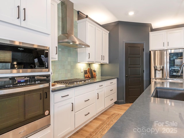 kitchen featuring wall chimney range hood, light wood-style flooring, white cabinets, stainless steel appliances, and a sink