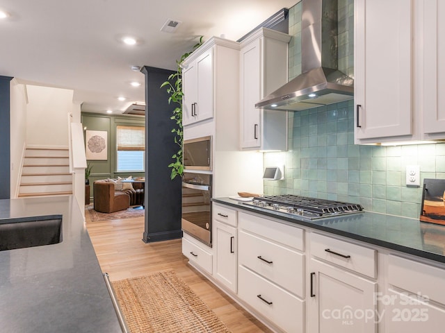 kitchen with dark countertops, wall chimney exhaust hood, wall oven, and stainless steel gas cooktop