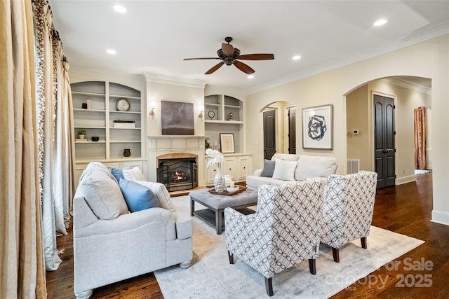 living room featuring crown molding, dark wood finished floors, recessed lighting, a warm lit fireplace, and arched walkways