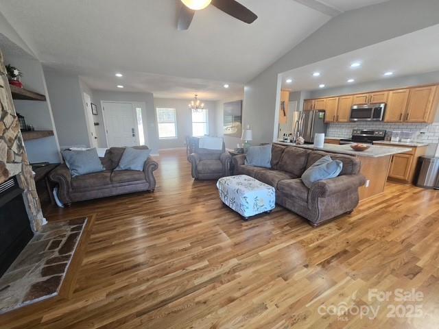 living room with light wood-type flooring, lofted ceiling, a stone fireplace, recessed lighting, and ceiling fan with notable chandelier