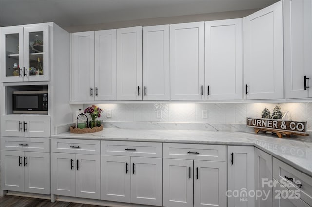 kitchen featuring stainless steel microwave, backsplash, glass insert cabinets, light stone counters, and white cabinetry