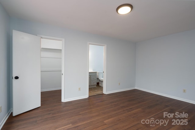 unfurnished bedroom with a spacious closet, baseboards, a closet, ensuite bathroom, and dark wood-style floors