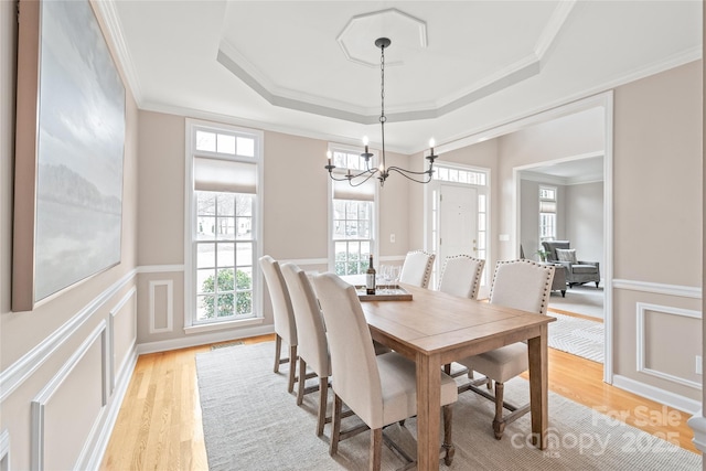 dining space featuring a tray ceiling, light wood-style floors, wainscoting, crown molding, and a decorative wall