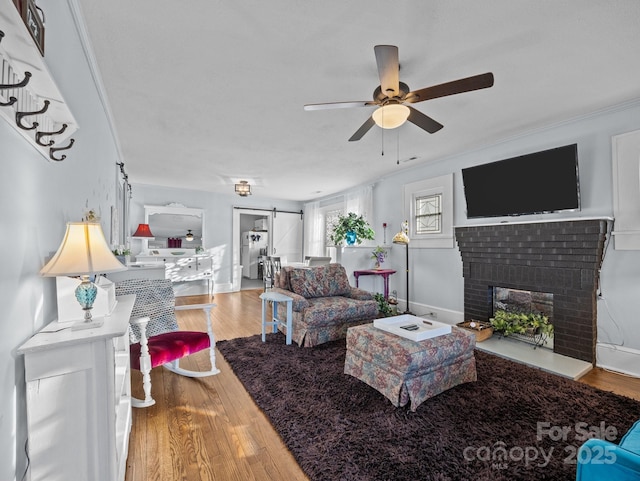 living area featuring a ceiling fan, wood finished floors, a barn door, baseboards, and a brick fireplace