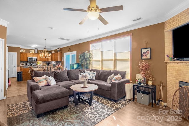 living area featuring light wood finished floors, visible vents, crown molding, ceiling fan with notable chandelier, and a fireplace