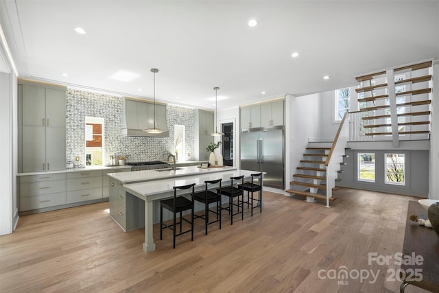 kitchen with a center island with sink, light wood finished floors, a sink, wall chimney range hood, and stainless steel built in refrigerator