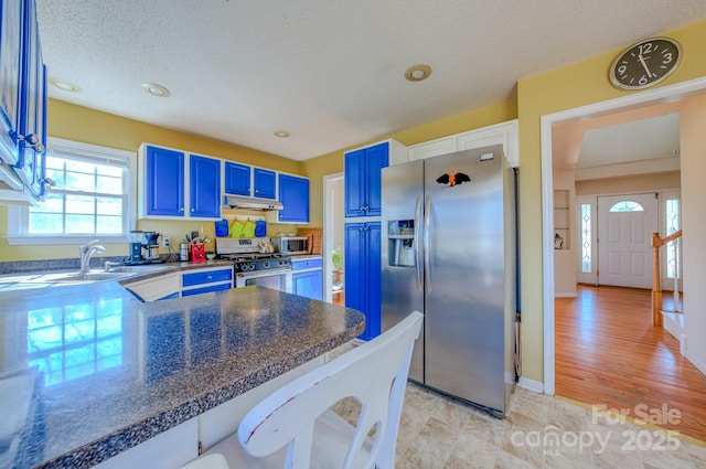 kitchen featuring dark countertops, blue cabinetry, under cabinet range hood, stainless steel appliances, and a sink