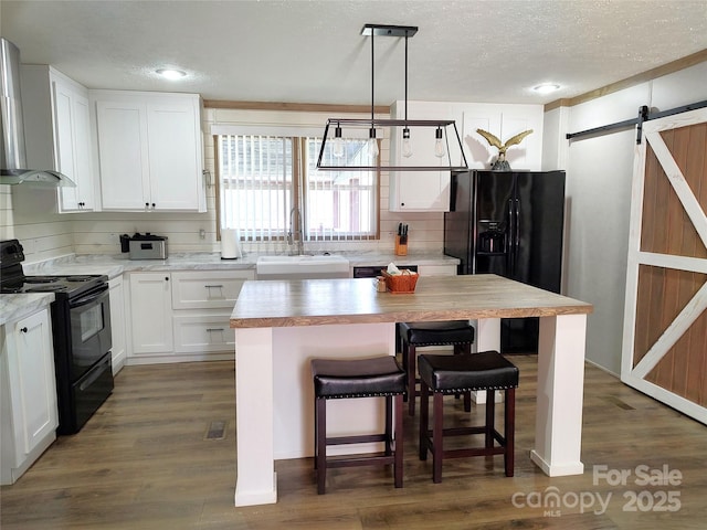 kitchen with a sink, black appliances, white cabinets, a barn door, and wall chimney range hood