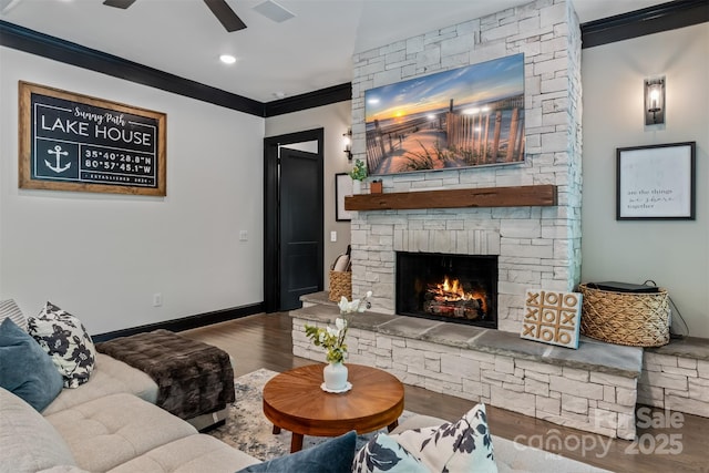 living room featuring wood finished floors, baseboards, ceiling fan, a stone fireplace, and crown molding