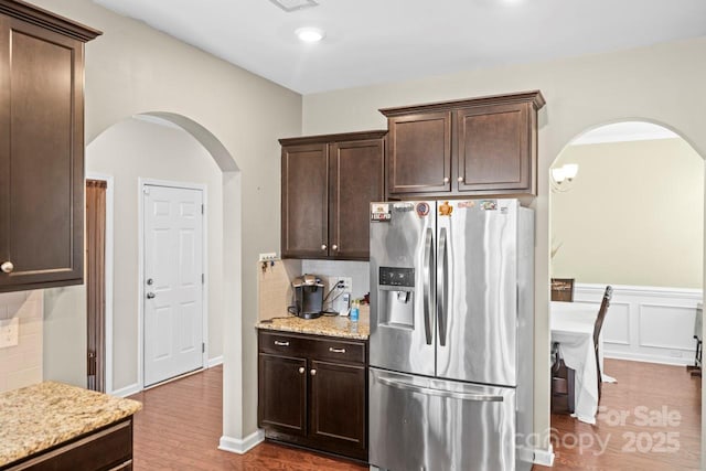 kitchen featuring light stone counters, wood finished floors, stainless steel fridge, arched walkways, and dark brown cabinets