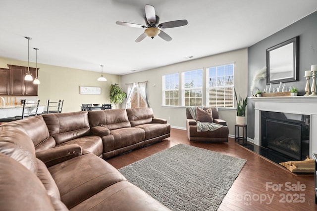 living area featuring visible vents, a ceiling fan, a glass covered fireplace, dark wood finished floors, and baseboards