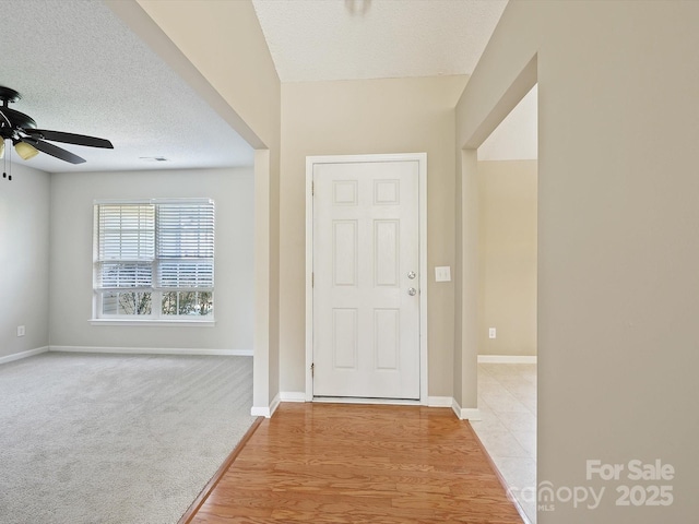 entryway featuring baseboards, light colored carpet, light wood-type flooring, a textured ceiling, and a ceiling fan