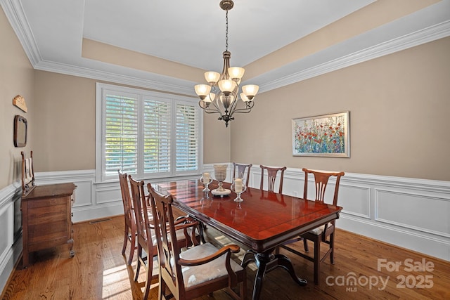 dining area featuring light wood-type flooring, a tray ceiling, ornamental molding, wainscoting, and a notable chandelier