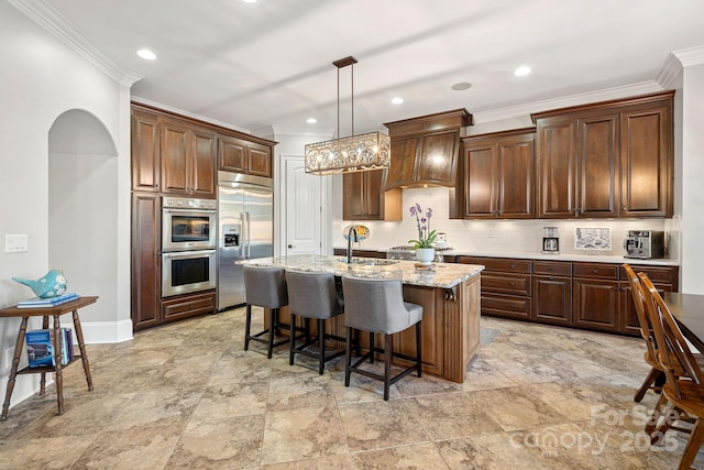 kitchen with tasteful backsplash, a sink, custom exhaust hood, stainless steel appliances, and a kitchen island with sink