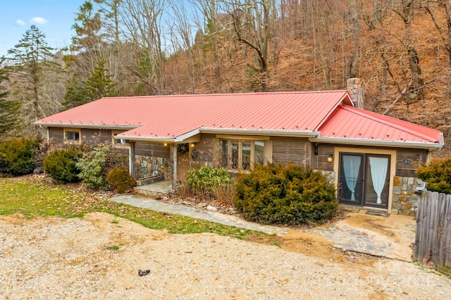 single story home featuring a chimney, stone siding, metal roof, and a view of trees