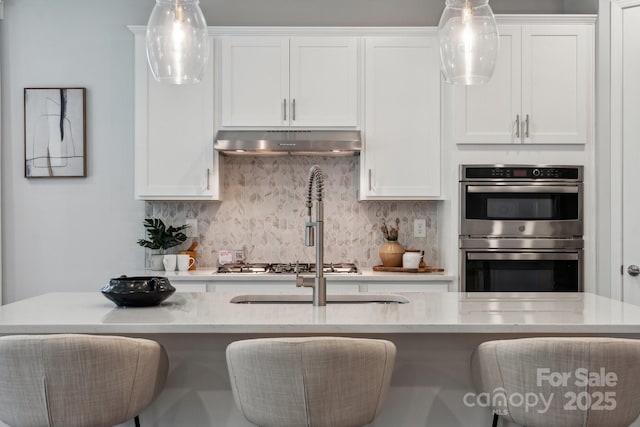 1439 Hubert Graham Way, Tega Cay, SC 29708 - kitchen featuring under cabinet range hood, double oven, decorative backsplash, white cabinets, and hanging light fixtures