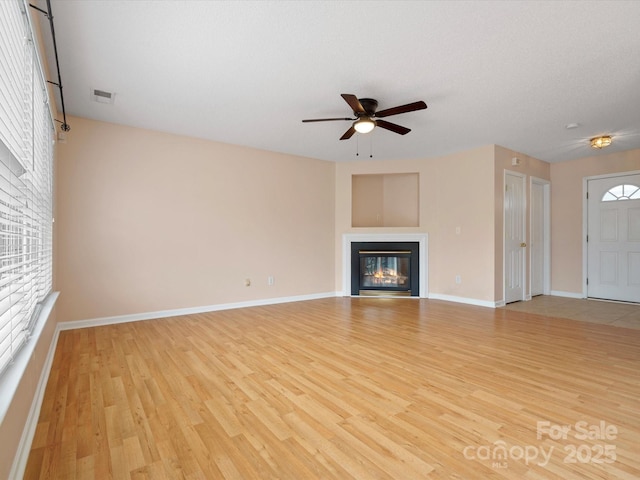 unfurnished living room featuring visible vents, baseboards, wood finished floors, a glass covered fireplace, and a ceiling fan