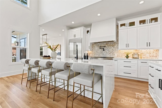 kitchen with stainless steel built in refrigerator, custom range hood, backsplash, light countertops, and an island with sink