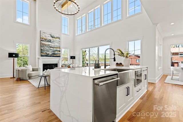 kitchen with light wood-style flooring, a towering ceiling, a glass covered fireplace, dishwasher, and light stone counters