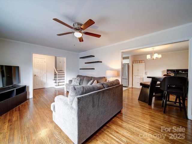 living room with ornamental molding, ceiling fan with notable chandelier, wood finished floors, stairway, and baseboards