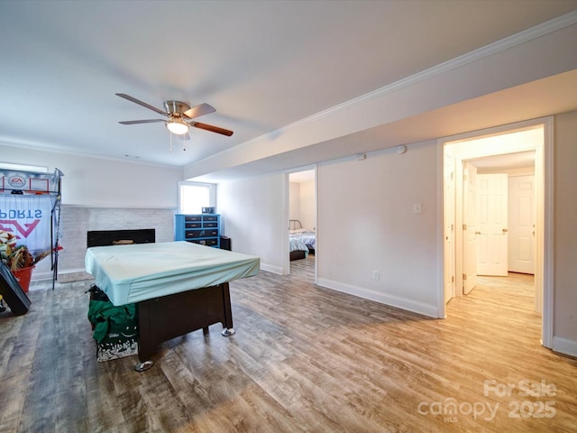 playroom featuring wood finished floors, baseboards, ceiling fan, crown molding, and a brick fireplace