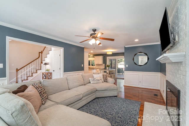 living area with wood finished floors, a wainscoted wall, a fireplace, ornamental molding, and ceiling fan