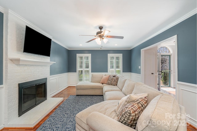 living area with a wainscoted wall, wood finished floors, crown molding, a brick fireplace, and ceiling fan