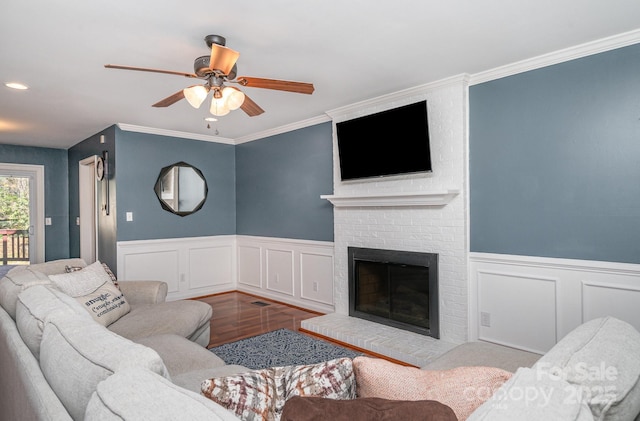 living area featuring a wainscoted wall, wood finished floors, crown molding, a brick fireplace, and ceiling fan