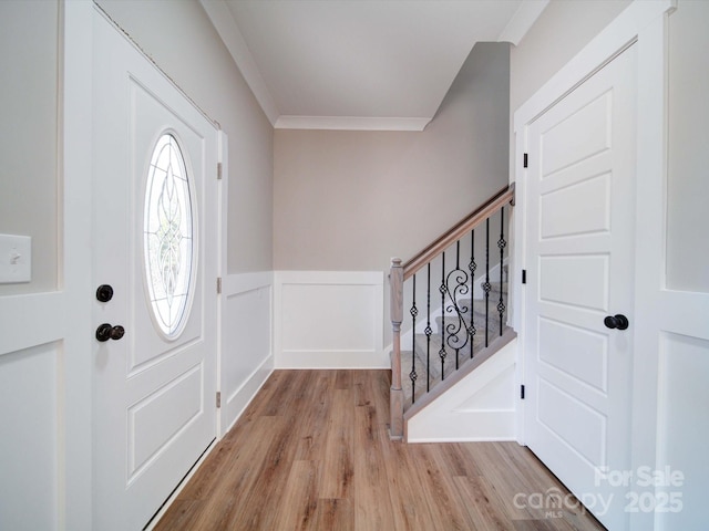 foyer featuring stairway, light wood-style floors, wainscoting, crown molding, and a decorative wall