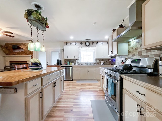 kitchen with wall chimney range hood, butcher block countertops, appliances with stainless steel finishes, a ceiling fan, and a sink