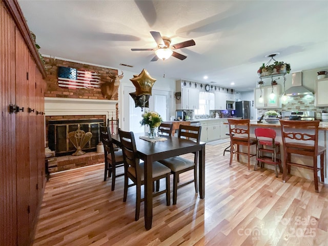 dining room featuring light wood-type flooring, a brick fireplace, ceiling fan, and visible vents