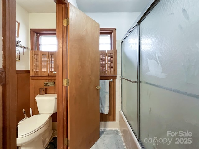 bathroom featuring toilet, a healthy amount of sunlight, wainscoting, and shower / bath combination with glass door