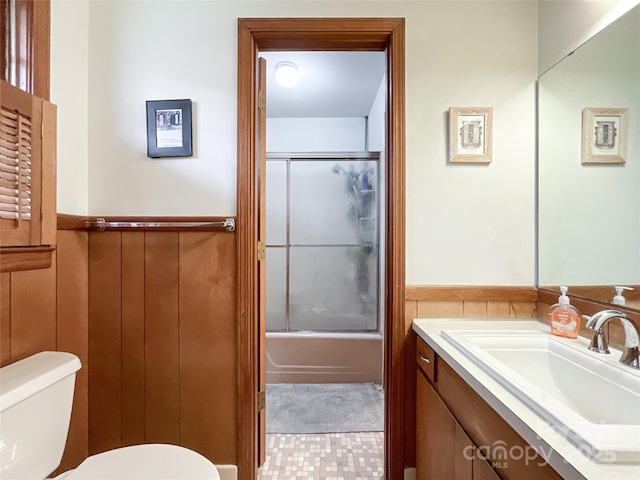 bathroom with a wainscoted wall, toilet, vanity, and bath / shower combo with glass door