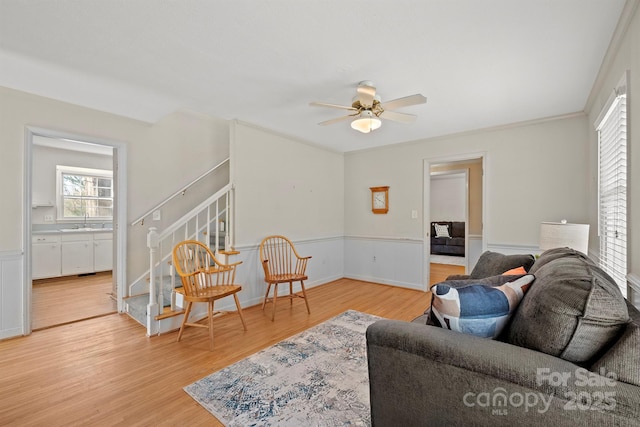 living room featuring light wood-type flooring, a wainscoted wall, ceiling fan, and stairs