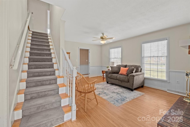 living room with a ceiling fan, stairway, wood finished floors, and a wainscoted wall