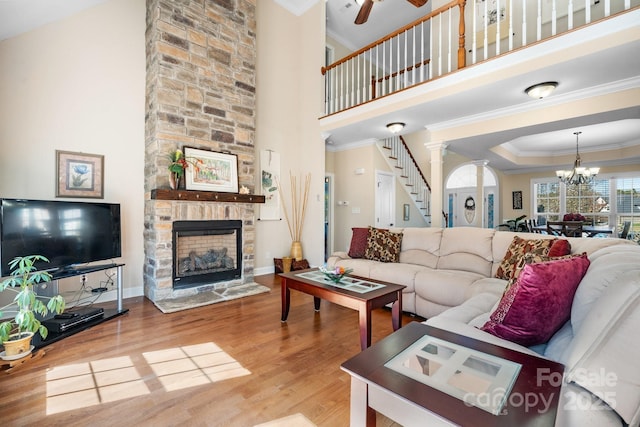 living room featuring ceiling fan with notable chandelier, wood finished floors, a stone fireplace, crown molding, and stairs