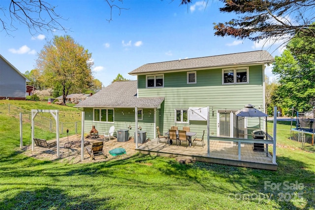 back of house featuring a trampoline, a fire pit, central AC unit, a wooden deck, and a lawn