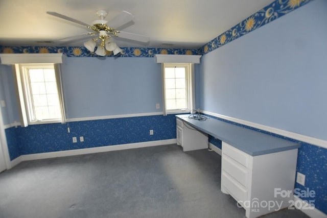 unfurnished office featuring baseboards, wallpapered walls, ceiling fan, built in desk, and dark colored carpet