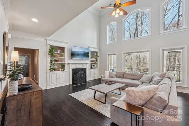 living area featuring a fireplace with flush hearth, ornamental molding, dark wood finished floors, baseboards, and ceiling fan