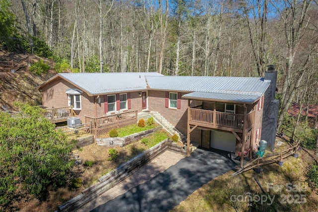 chalet / cabin with central air condition unit, driveway, a view of trees, brick siding, and a chimney